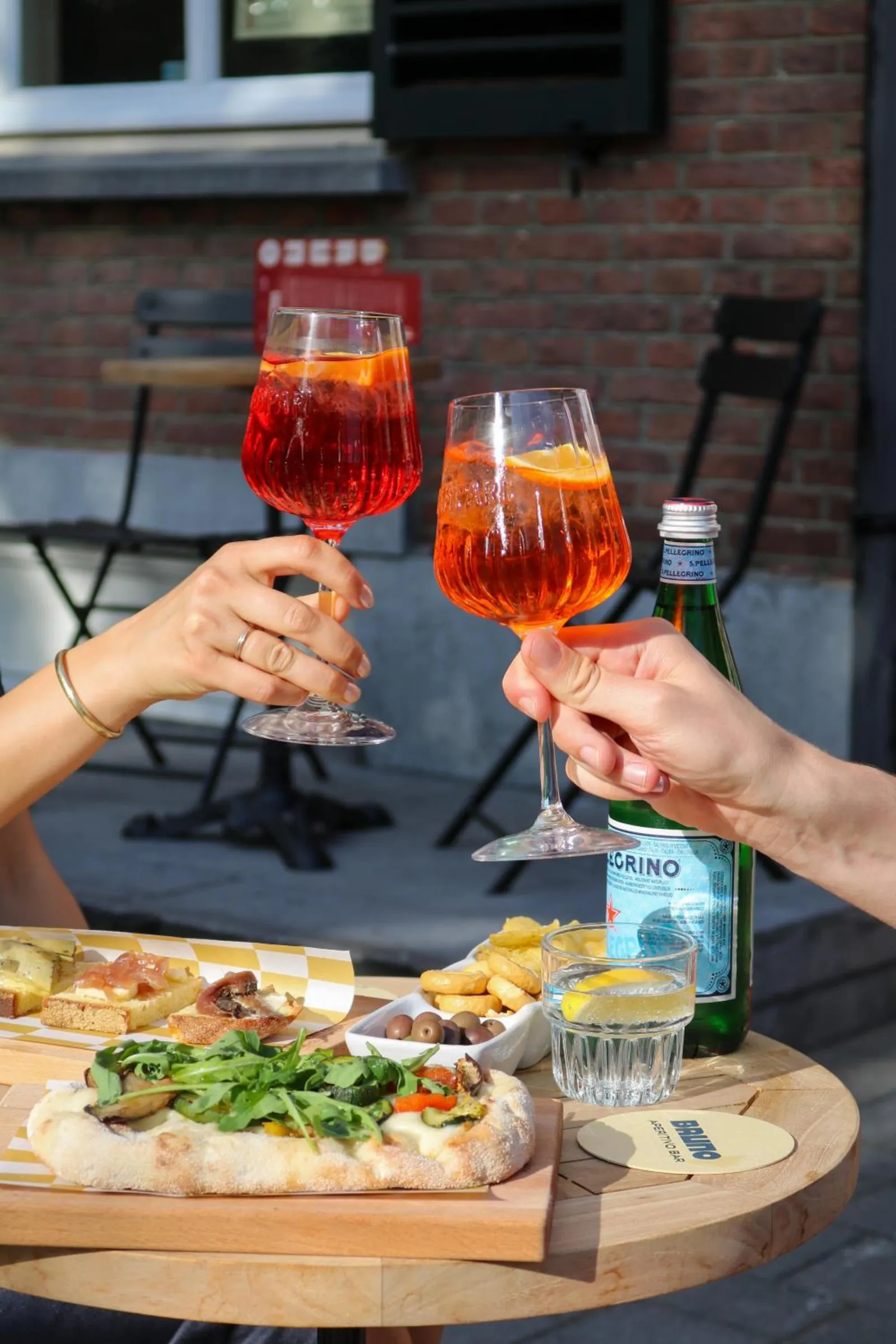 Two glasses of spritz raised in a toast over pizza and snacks on the terrace
