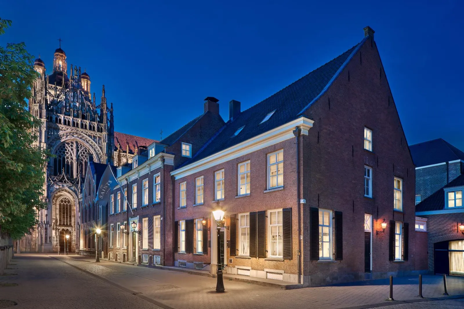 The brick facade of The Saints Den Bosch on the Parade at blue hour, with the illuminated St John's Cathedral alongside