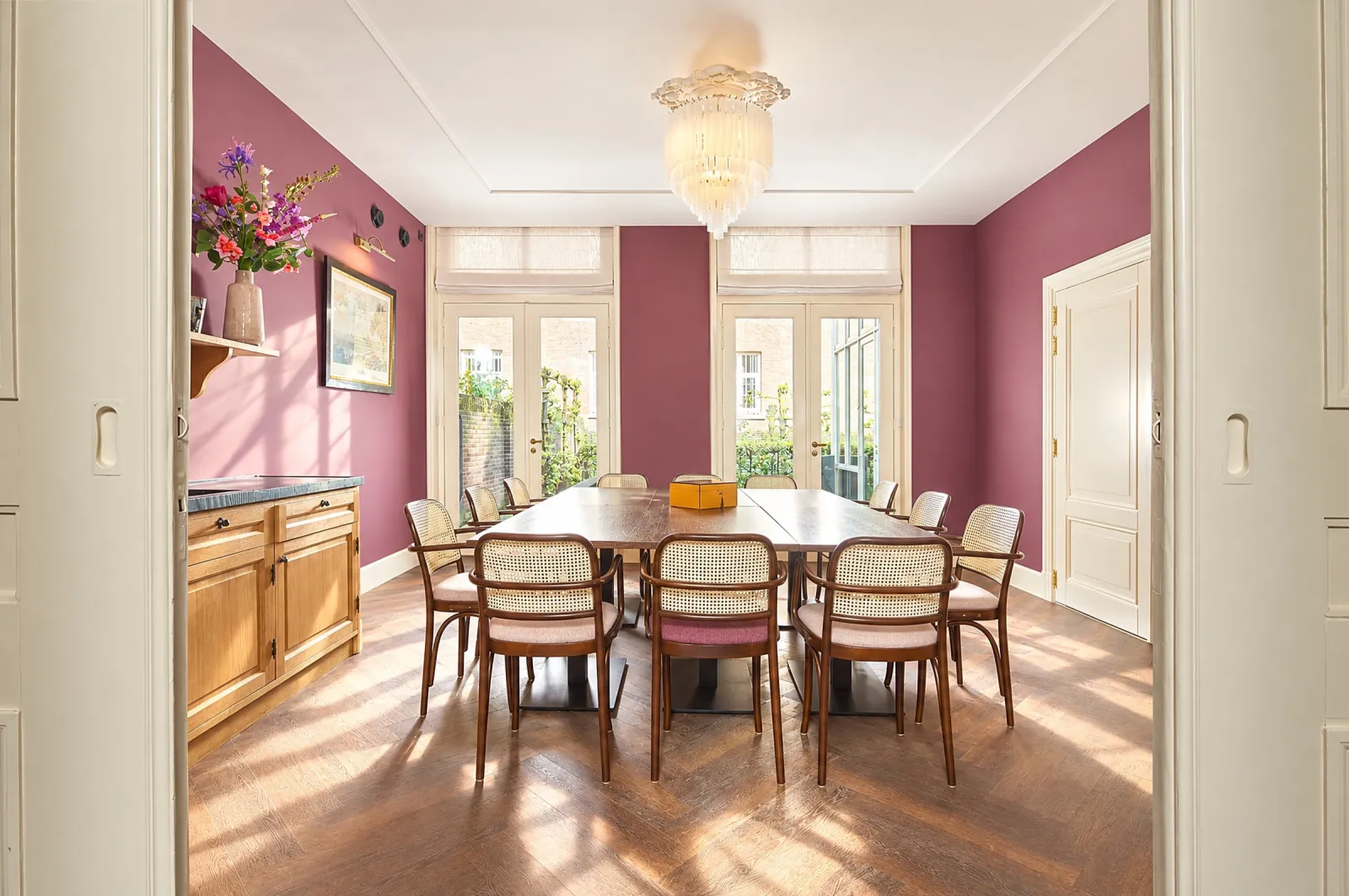 Besloten eetkamer met roze wanden, lange tafel, kroonluchter en tuindeuren